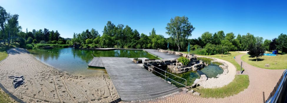 Bild des Naturerlebnisbades mit seinen Teichen, dem Naturstrand und dem Waldambiente zwischen den schattenspendenden Bäumen.