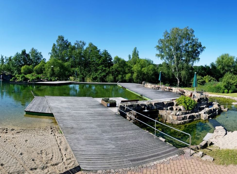 Bild des Naturerlebnisbades mit seinen Teichen, dem Naturstrand und dem Waldambiente zwischen den schattenspendenden Bäumen.